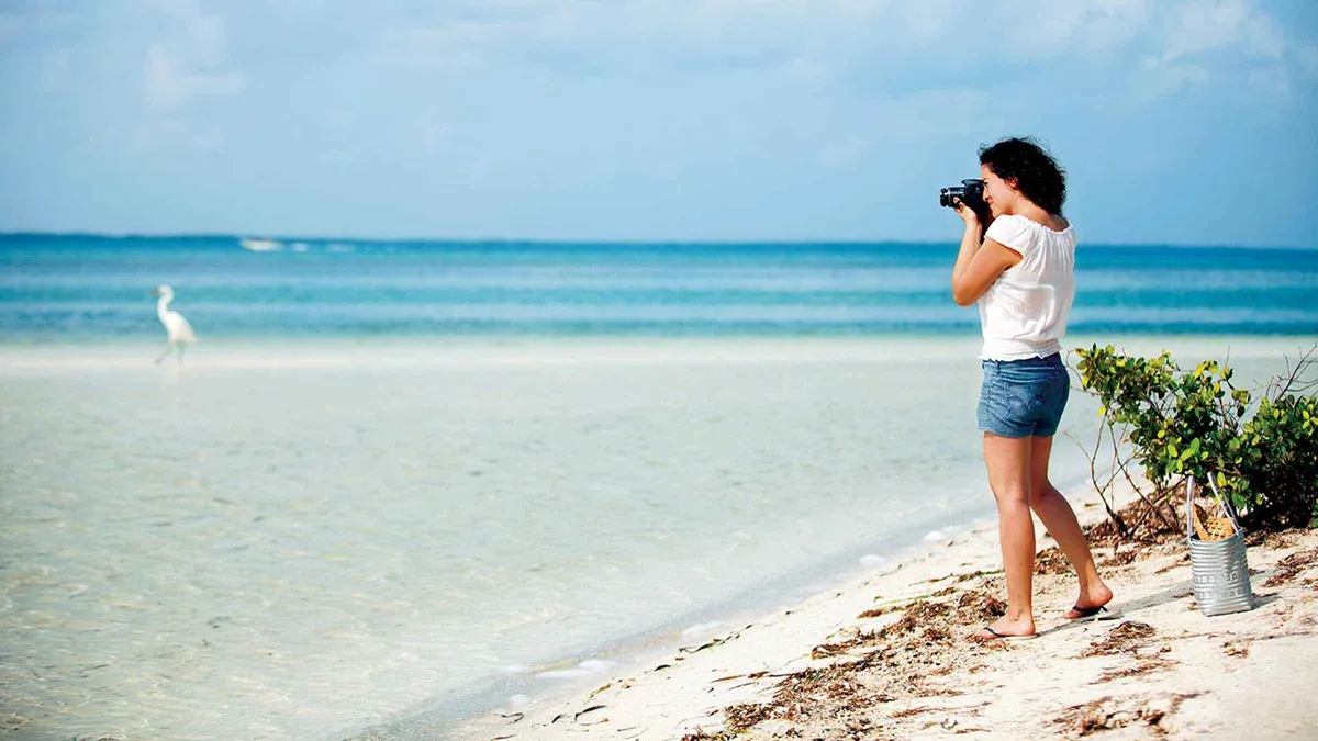 A woman taking photos on a white sand beach beside shallow turquoise water